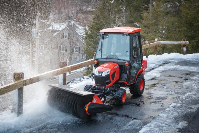 Kubota BX2680 tractor with enclosed cab and snow broom attachment clearing a snowy roadway